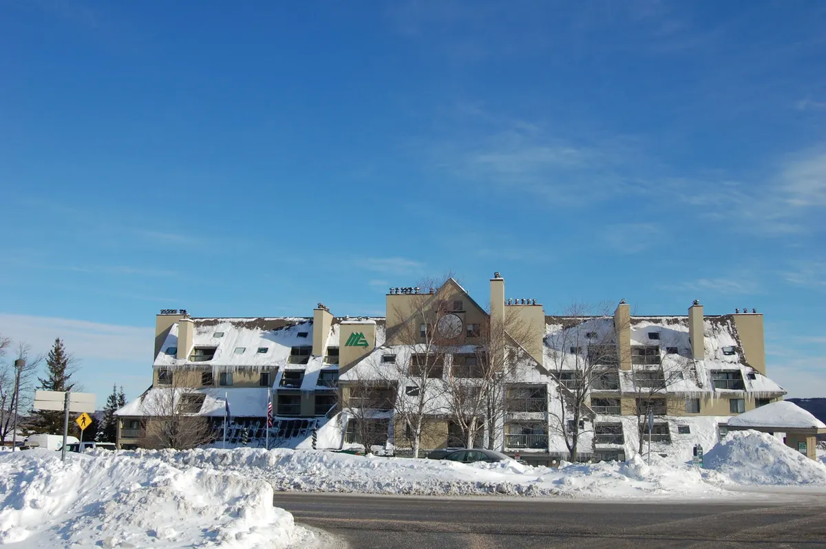 A snow-covered building is nestled among tall trees beneath a clear blue sky. The image evokes a cozy, adventurous winter atmosphere, suggesting proximity to skiing and winter sports while retaining a sense of comfort and retreat.