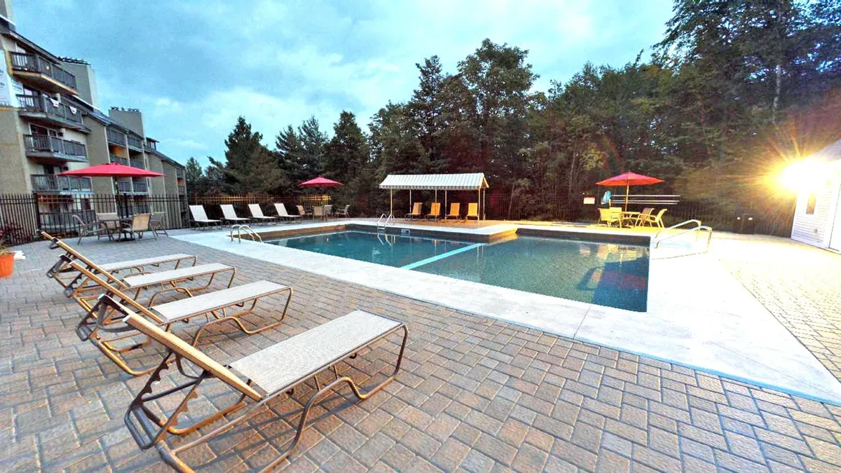 An inviting outdoor pool surrounded by lounge chairs, cabanas, and cheerful red umbrellas. The setting is bathed in warm light, suggesting a laid-back and social atmosphere perfect for sunny days and summer relaxation.