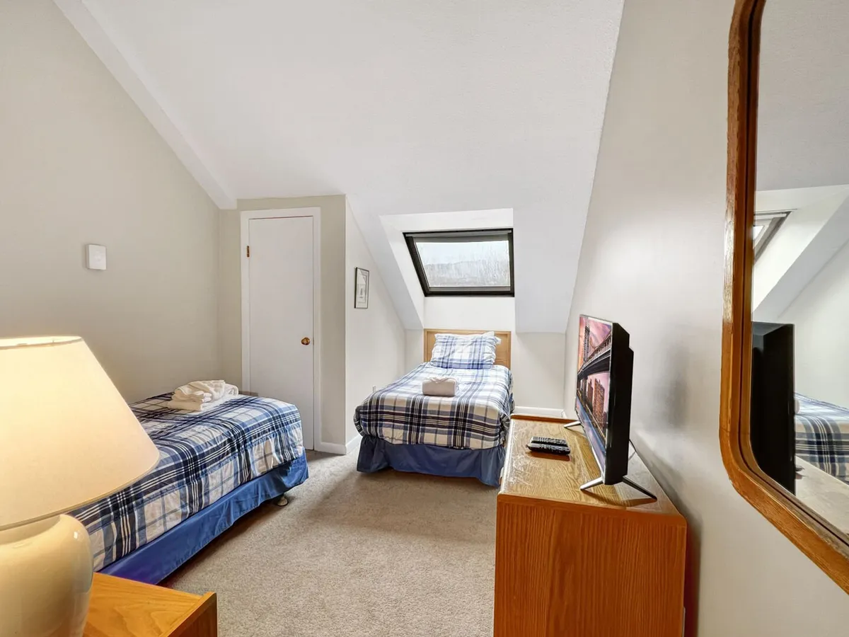 A warmly lit bedroom with two beds dressed in plaid covers, a window streaming in daylight, a decorative mirror, and a nightstand with a lamp. The relaxed mood is furthered by soft furnishings and thoughtful touches, making it an inviting retreat for restful nights.