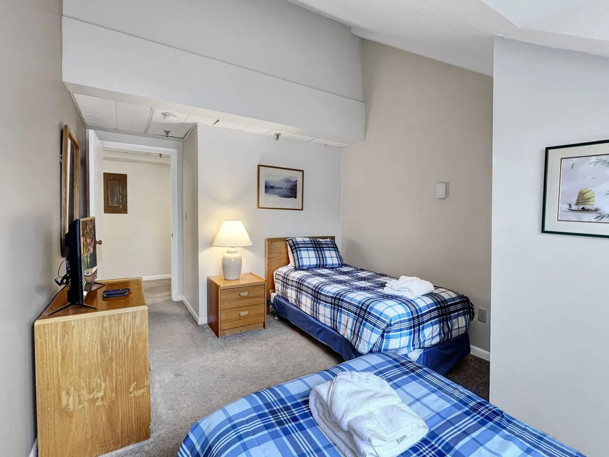 This image features the second bedroom, with two twin beds adorned with plaid bedding, a nightstand with a lamp between them, a flat-screen TV on a wooden dresser, and framed artwork on the wall. The ample window lets in natural light, creating a cozy and inviting space ideal for families or friends sharing the room.