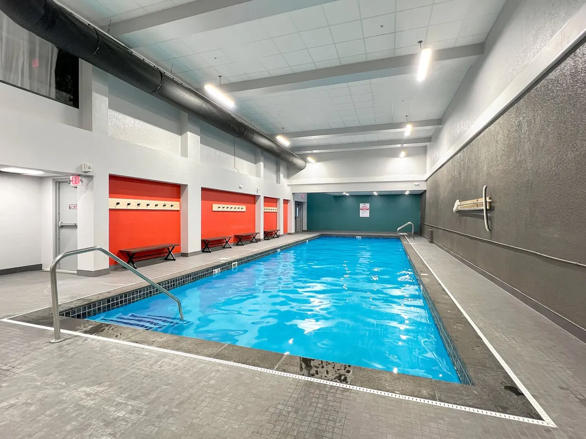 A bright and energetic indoor swimming pool area featuring vivid blue water, red accent walls, and benches along the pool perimeter. The space is well-lit and ideal for year-round swimming, socializing, and fitness activities.