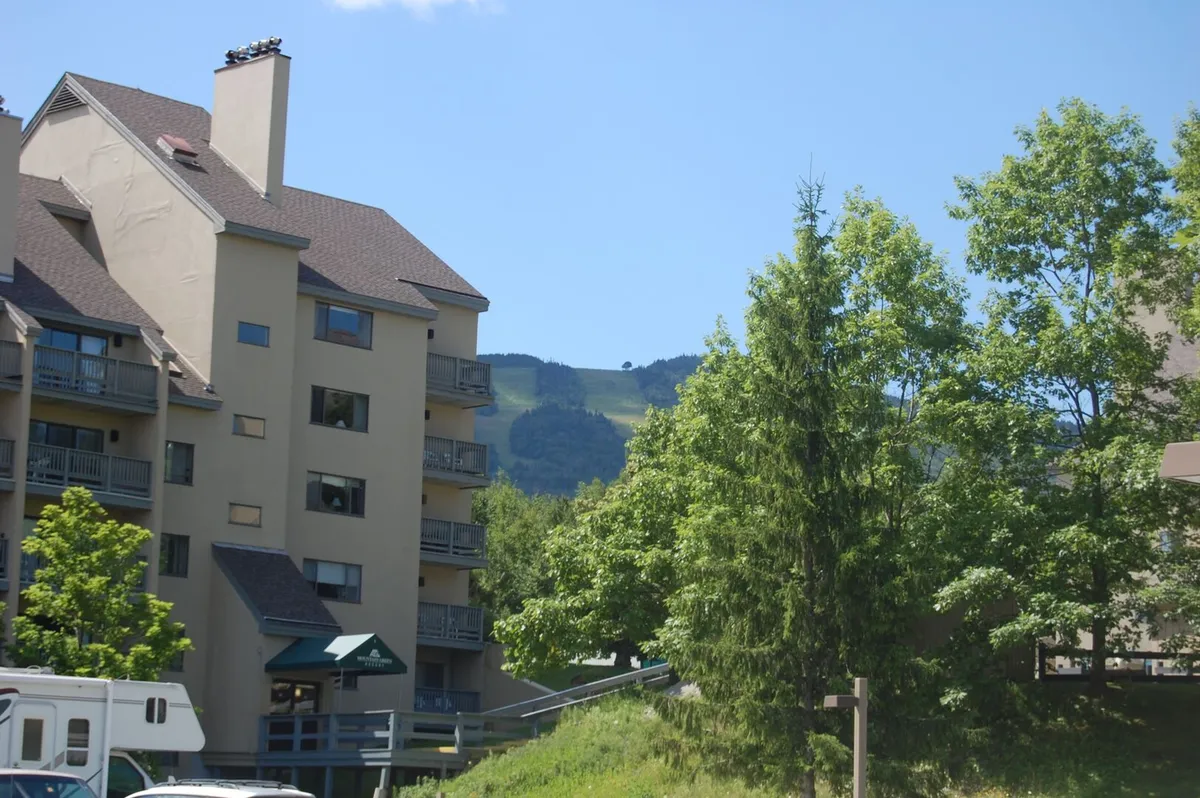 A striking exterior shot of the building facade framed by lush green trees and a stunning mountain backdrop. The scene is tranquil, showcasing access to nature trails and the beauty that surrounds the property.