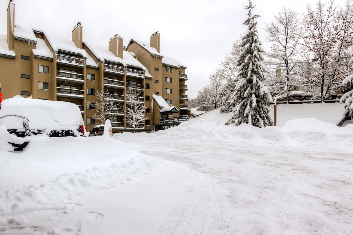 A snow-covered apartment building is surrounded by snow-laden trees and parked vehicles. The tranquil setting suggests easy access to winter sports and immediately places the viewer in a classic ski resort environment. The scene captures the heart of a ski destination, showing the proximity of lodging to snowy action and the welcoming winter atmosphere.
