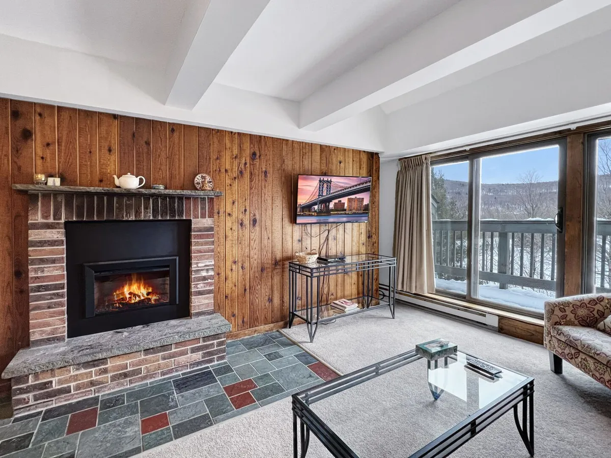 A warm, inviting living room featuring a large window framed by a wooden accent wall, a fireplace, a TV, and a coffee table. The ample seating and expansive glass doors allow for appreciation of mountain vistas from within the cozy interior.